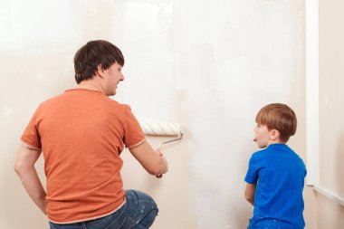 Genç bir aile duvarları boyuyor. Baba ve oğul duvar boyuyor. Mutlu aile yeni evlerini yeniliyor. Baba oğluna tekerlekle nasıl duvar boyadığını gösteriyor..