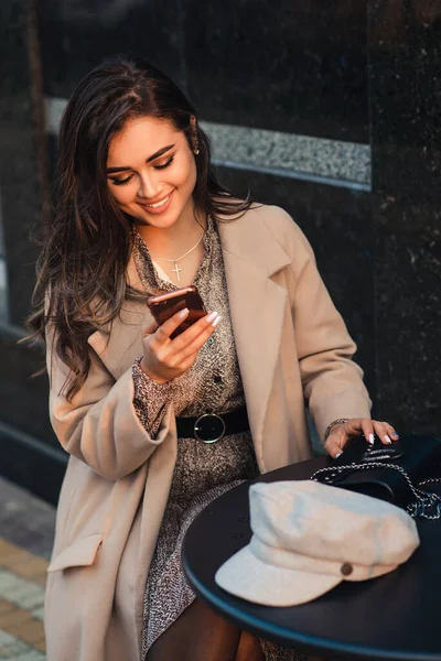 Women fashion. Woman with perfect makeup and hairstyle. Seductive girl at coffee shop outside. Technology and lifestyle. Fashionable girl with mobile phone sitting at cafe outdoors. City lifestyle.