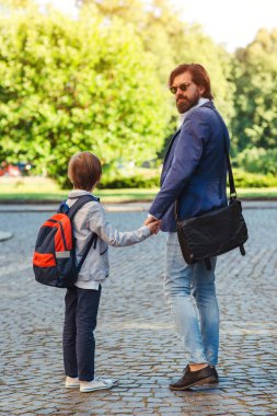 Okula giderken sırt çantalı bir çocuk babasının elini tutuyor. Okula dönelim. Baba oğlunu okula götürür. Ebeveynlik, ilişki, çocukluk.