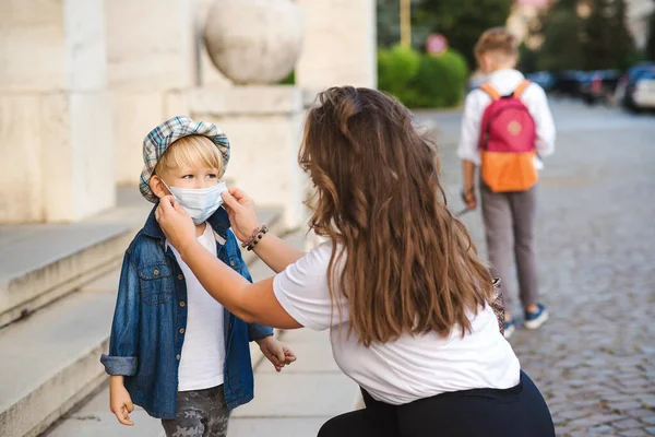 Mother puts a safety mask on her son. Cute boy with a backpack outdoors. Back to school concept. Medical mask to prevent coronavirus. Coronavirus quarantine.
