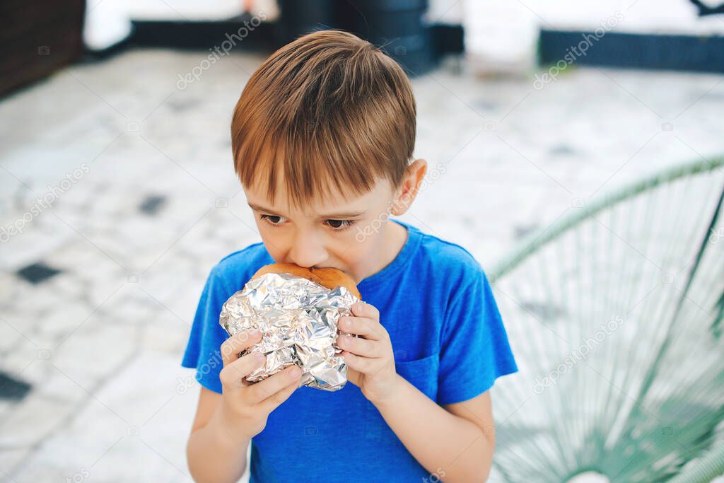 Hungry boy eating a burger at outdoors cafe. Cute child eating fast food. Childhood, unhealthy food concept. Junky food. Lifestyle and people