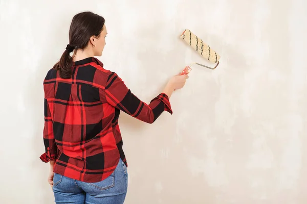 Woman paints wall with paint roller. Skillful and professional painter. Home renovation concept. Young woman primed wall. Female painting wall in the room, back view.