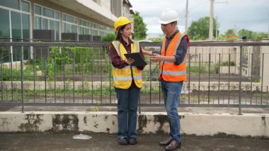Bir projeyi inceleyen iki inşaat mühendisi, Asyalı bir kadın mimar ve bir erkek müteahhit, inşaat alanındaki bir binanın etrafında yürürken ellerinde tabletler ve panolar tutuyorlardı..