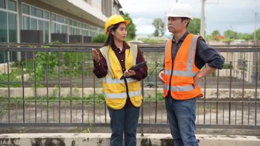 Bir projeyi inceleyen iki inşaat mühendisi, Asyalı bir kadın mimar ve bir erkek müteahhit, inşaat alanındaki bir binanın etrafında yürürken ellerinde tabletler ve panolar tutuyorlardı..