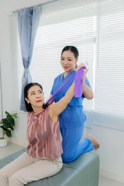 Asyalı fizyoterapist hastanın yatakta elastik bantla egzersiz yapmasına yardım ediyor. Klinikte orta yaşlı Asyalı kadınların elastik bantla egzersiz yapmasına yardım etmek..