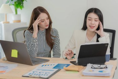 Two Asian businesswomen working with laptop and tablet computers discussing documents on desk, smiling colleagues discussing new project, business