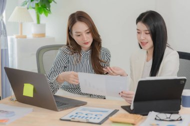 Two Asian businesswomen working with laptop and tablet computers discussing documents on desk, smiling colleagues discussing new project, business
