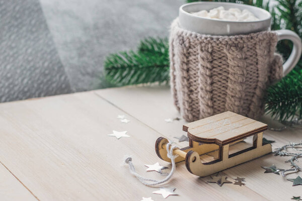 White cup for cocoa in beige knitted clothes. Santa Claus sleigh on a white wooden background with christmas tree space for text