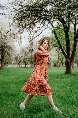 Young beautiful blonde woman in blooming garden. Spring trees in bloom. Orange dress and straw hat.