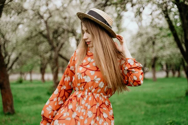 Young beautiful blonde woman in blooming garden. Spring trees in bloom. Orange dress and straw hat. — Stock Photo, Image
