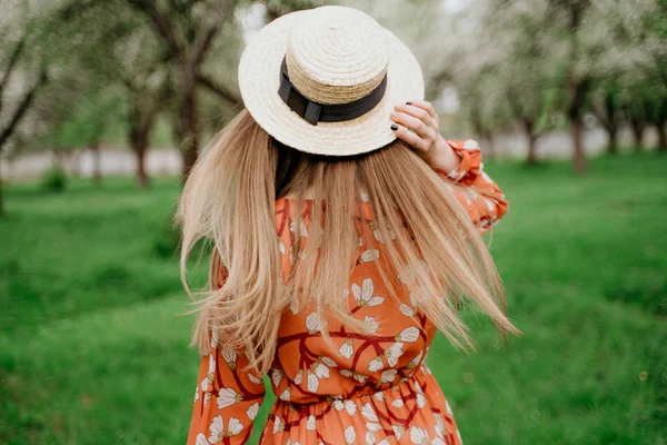 Young beautiful blonde woman in blooming garden. Spring trees in bloom. Orange dress and straw hat. — Stock Photo, Image