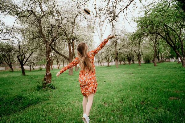 Young beautiful blonde woman in blooming garden. Spring trees in bloom. Orange dress and straw hat. — Stock Photo, Image