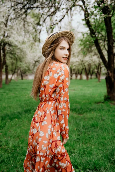 Young beautiful blonde woman in blooming garden. Spring trees in bloom. Orange dress and straw hat. — Stock Photo, Image