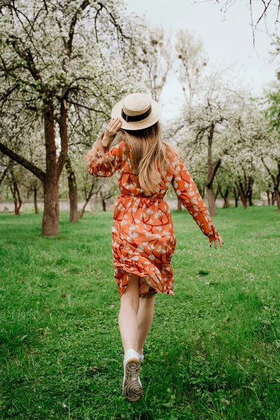 Young beautiful blonde woman in blooming garden. Spring trees in bloom. Orange dress and straw hat. — Stock Photo, Image