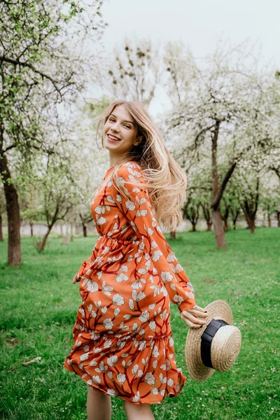 Young beautiful blonde woman in blooming garden. Spring trees in bloom. Orange dress and straw hat. — Stock Photo, Image