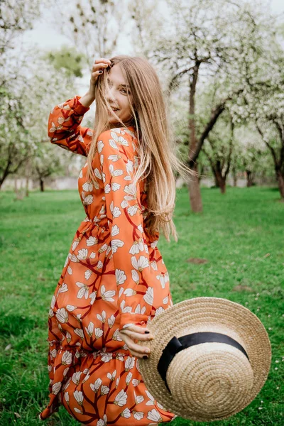 Young beautiful blonde woman in blooming garden. Spring trees in bloom. Orange dress and straw hat. — Stock Photo, Image