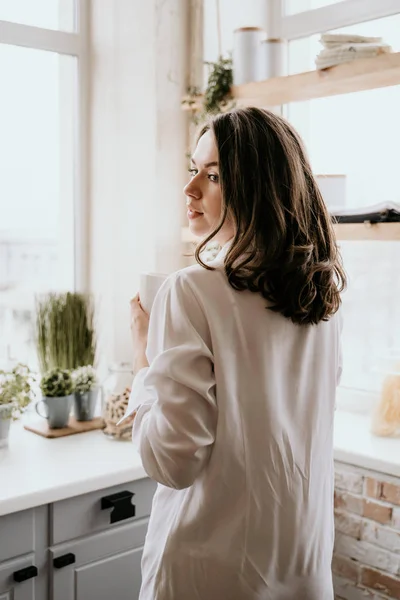 Mujer exitosa y segura en una pausa para tomar café por la mañana, descansando y disfrutando de la bebida. Chica joven en bragas blancas y camisa larga con hombro desnudo y el pecho de pie en la cocina de la casa — Foto de Stock