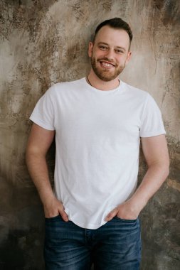 Adam kot pantolon ve dokusal bir arka plan üzerinde beyaz bir t-shirt gülümsüyor. Kızıl saç ve sakal.