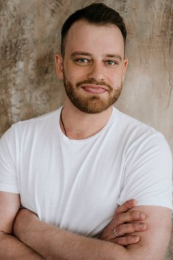 Adam kot pantolon ve dokusal bir arka plan üzerinde beyaz bir t-shirt gülümsüyor. Kızıl saç ve sakal.