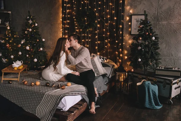 Un par en la cama del dormitorio. Interior oscuro. Año Nuevo y Navidad. Abrazos y besos. Amor. Jersey blanco y calcetines altos. Reunión romántica. Amantes de las citas. — Foto de Stock