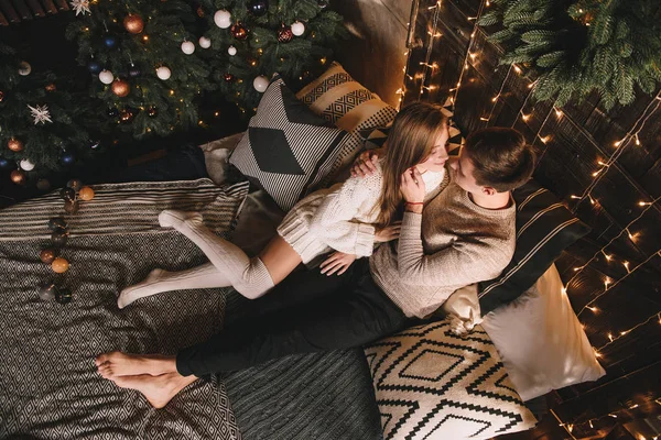 Paar auf dem Bett im Schlafzimmer. Dunkles Interieur. Neujahr und Weihnachten. Umarmungen und Küsse. Liebe. Weißer Pullover und hohe Socken. Romantisches Treffen. Ein Date. — Stockfoto