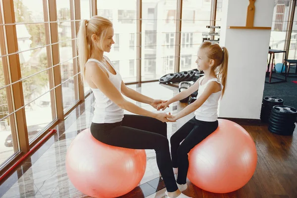 Mom and Daughter are doing yoga. Family in a gym. Little girl with mother in a white t-shirts and black leggings. Girls with red fitball