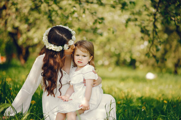 Beautiful young pregnant girl in a long white dress and wreath on her head playing in a sunny summer park between the trees with her little daughter