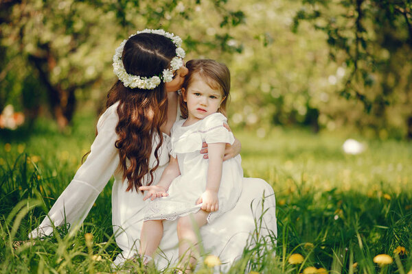 Beautiful young pregnant girl in a long white dress and wreath on her head playing in a sunny summer park between the trees with her little daughter