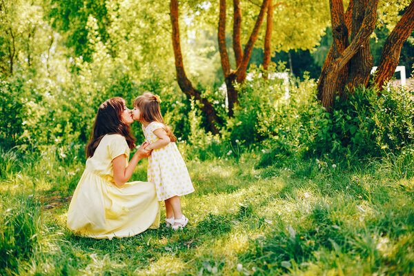 In a summer solar park near green trees, mom walks in a yellow dress and her little pretty girl