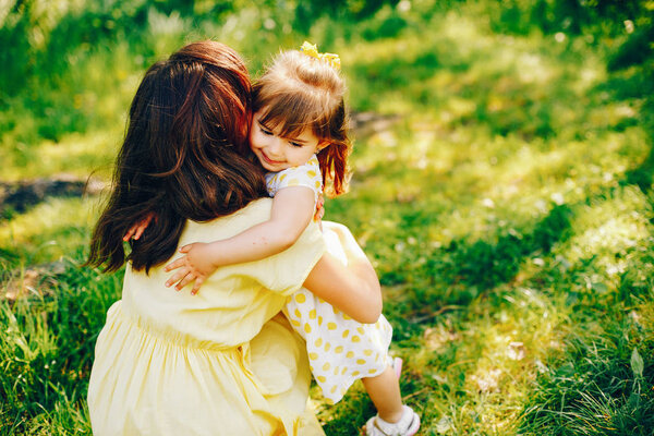 In a summer solar park near green trees, mom walks in a yellow dress and her little pretty girl