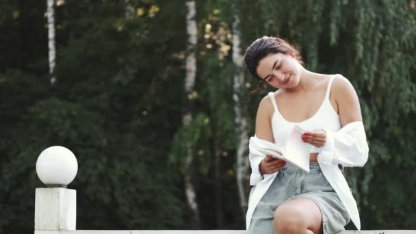 Joven bonita mujer sonriendo y leyendo libro en el parque verde — Vídeo de stock