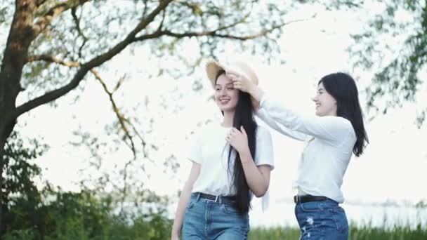 Two girlfriends, one wearing a straw hat at the lakeside smiling — Stock Video