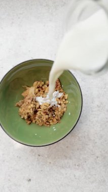 Yavaş çekimde süt, açık renkli mutfak tezgahında gevrek granola parçalarıyla dolu yeşil kaseye dökülüyor.
