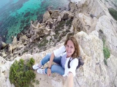 Bir uçurum ve güzel bir deniz manzarası arka planda bir selfie alarak bir sırt çantası ile genç bir kadın oturuyor. Güler yüz.