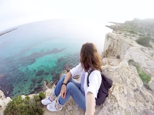 一个带着背包的年轻女子坐在海面上的悬崖上 看着地平线 自拍在风景秀丽的背景下 — 图库视频影像