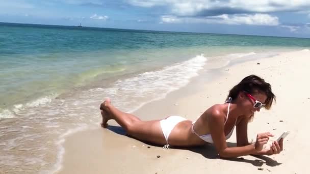 穿着比基尼的年轻漂亮女子躺在沙滩上 手里拿着一把智能手机 — 图库视频影像