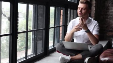 Genç bir adam ve bir pencere üzerinde oturur bir dizüstü bilgisayar ve bir smartphone kullanarak çalışır.