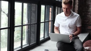 Genç bir adam ve bir pencere üzerinde oturur bir dizüstü bilgisayar ve bir smartphone kullanarak çalışır. Modern iş adamı.