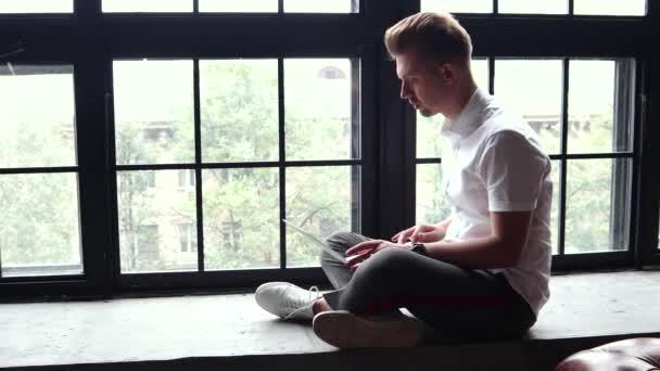 Un joven se sienta en un alféizar de la ventana y trabaja con un ordenador portátil y un teléfono inteligente . — Vídeo de stock