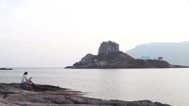 En ung kvinna sitter med sin smartphone på rocky shore vid havet. — Stockvideo