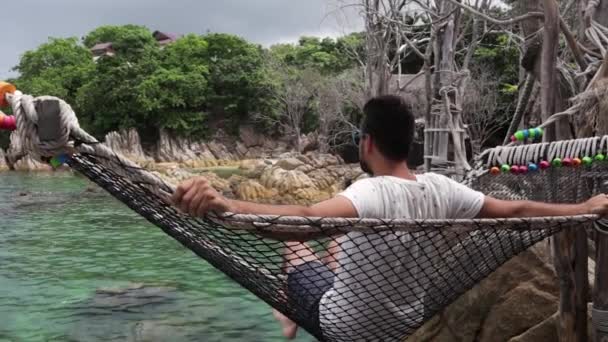 Joven hombre guapo se balancea en la hamaca con una vista al mar . — Vídeo de stock