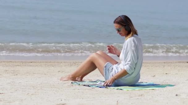 Een jonge vrouw is luisteren naar muziek op het strand op een zonnige dag. — Stockvideo