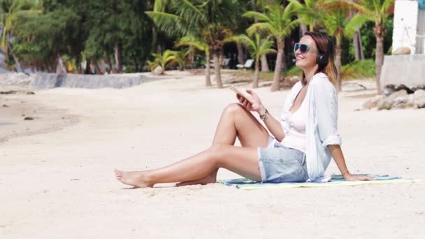 Een jonge vrouw is luisteren naar muziek op het strand op een zonnige dag. — Stockvideo