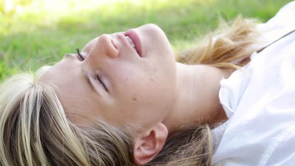 Jonge blonde vrouw met mooie blauwe ogen ligt op het gras op het Park. — Stockvideo