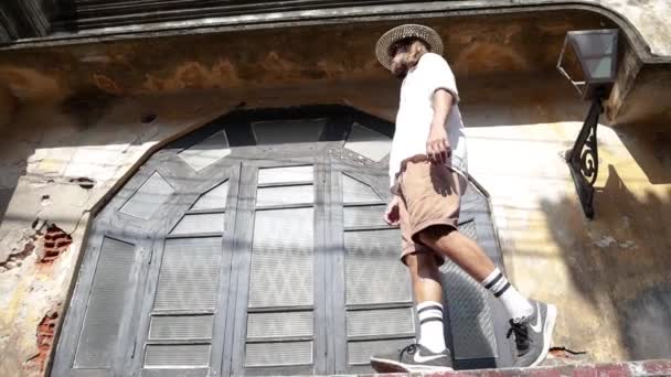 麦わら帽子とサングラスをかけた若い男が 古い歴史的な家の狭い縁石の上でバランスをとろうとしている — ストック動画