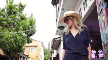 Hasır şapkalı ve modaya uygun güneş gözlükleri ile genç ve güzel sarışın bir kadın sokakta duruyor.