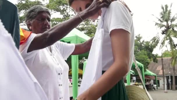 Een jonge blanke vrouw is op de Sri Lanka lokale markt proberen op een shirt. — Stockvideo