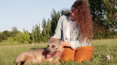 Uzun kıvırcık saçlı mutlu genç beyaz kadın yaz aylarında parkta köpeği ile iyi vakit geçiriyor.
