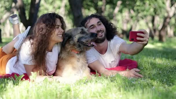Un jeune couple joyeux prenant un selfie au parc avec leur chien baisé — Video