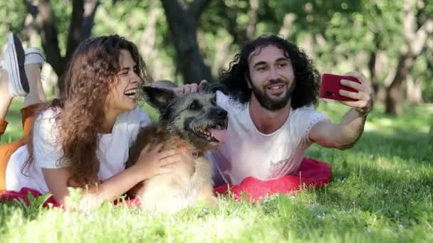 Молодая веселая пара делает селфи в парке со своей лохматой собакой — стоковое видео